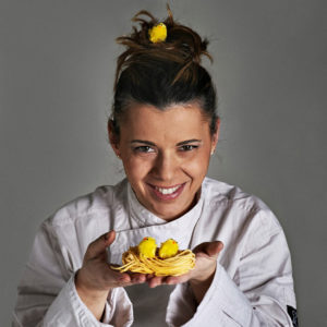 Chef Iside De Cesare Ambasciatrice del Gusto Doc Italy per la Calabria Cuoca, pasticcera, moglie e mamma. Iside De Cesare è tutto questo. Donna multi tasking dalle mille sfaccettature, incarna il modello femminile dell’era moderna capace di conciliare il lavoro con la famiglia, distribuendo tempo ed energia sia all’uno che all’altra. Ma chi è Iside De Cesare? Iside Maria De Cesare, classe 1973 nasce a Roma, dove frequenta la facoltà di ingegneria dopo aver conseguito la maturità scientifica. Durante gli studi si diletta in cucina prima in un ristorante stagionale aperto insieme alle sorelle e successivamente consolida la propria esperienza in altre attività ristorative. Si appassiona così tanto alla cucina che decide di interrompere l’università per dedicare le proprie energie al mondo della ristorazione. Comincia il suo iter esplorativo tra il ristorante Agata e Romeo a Roma, le Colline Ciociare di Salvatore Tassa per approdare poi alla Pergola del Rome Cavalieri. Dopo due anni alla corte di Heinz Beck va in Romagna alla Frasca Di Castrocaro Terme e, dopo altri due anni come chef in varie strutture e alberghi, dal 2005 gestisce ed è cuoca, insieme al marito Romano Gordini, il ristorante La Parolina premiato con la stella Michelin nel 2009. La Parolina si trova a Trevinano, una piccola frazione del comune di Acquapendente in provincia di Viterbo, terra di confine tra le regioni, Lazio Umbria e Toscana. Un ristorante stellato dall’ambiente famigliare dove propone una cucina semplice, riconoscibile e di gusto: “Ho da sempre un grande senso della famiglia e quando cucino immagino sempre di farlo per il pranzo della domenica, quando tutti sono riuniti intorno al tavolo in un clima di festa e gioia. La tavola è convivialità e io cerco di darla anche attraverso i miei piatti.” Non si risparmia Iside che in questi anni, ben 27, ha saputo cogliere il meglio da ogni cosa che la vita le offriva, a partire dal diventare madre che per lei ha significato imparare a nutrire il commensale, ad andare oltre l’estetica e la tecnica fine a sè stessa: “Viviamo in un territorio ricchissimo che offre un incredibile varietà di materie prime e di ricette della tradizione. In questi anni è diventato sempre più stretto il mio rapporto con i fornitori locali da cui attingo per le materie prime. Ho riscoperto, per esempio, il pesce di lago e i legumi, così come utilizzo molto la cacciagione nei mesi invernali dove la mia cucina diventa più strutturata. Noi facciamo ancora la pasta fatta in casa con il mattarello e le conserve di pomodoro come quando ero bambina. Ho imparato a usare i prodotti locali quando ci sono ma anche a conservarli quando sono alla massima maturazione e usarli poi nei mesi dove non posso trovarli freschi, e buoni”. Non si fa molte domande Iside, lavora e si gode i suoi bambini, che le hanno insegnato tanto, vive la vita con entusiasmo e passione, cercando di fare sempre meglio, cosa che le riesca anche abbastanza bene visto che il ristorante La Parolina è stato premiato con la stella Michelin nel 2008: “La stella Michelin per me è stata importante, è una cosa di cui vado fiera e di cui sono rispettosa. Ma la soddisfazione più grande per me rimane la felicità dei clienti con cui mi confronto ogni giorno”. Foto di Marco Varoli Foto di Marco Varoli Non è un caso che Iside faccia parte dell’Atelierdes Grandes Dames, network creato da Veuve Clicquot che accoglie donne talentuose – come fu la fondatrice della Maison di Champagne che rimase vedova a 27 anni e prese in mano le redini della cantina facendone uno dei marchi più famosi del mondo – e celebra i talenti femminili dell’alta ristorazione, come la nostra piccola chef dai grandi occhi azzurri. Il ristorante si affaccia sulla Val d’Orca e dalle vetrate si gode di una vista mozzafiato sul Monte Amiata, mentre nella bella stagione di mangiare all’aperto in giardino o sulla terrazza. Per chi volesse staccare la spina dalla città c’è la possibilità di soggiornare in comode e spaziose stanze e gustare di una colazione curata da Iside “Per me la colazione è sacra: iniziare la giornata con il sorriso e tante leccornie è il modo migliore per affrontarla” Iside è tutta determinazione e sorrisi, e niente scorciatoie. Potresti mai scegliere un posto tanto appartato se vuoi la vita facile? Giammai. Meglio sapere che chi ti viene a trovare non capita per caso. Che poi: ora che c’è la stella, il libro delle prenotazioni è sempre fitto. Ma quando Iside e Romano si sono messi in proprio, 6 anni e passa fa, era dura. Un po’ li ha sorretti l’amore – quello romantico e quello per la cucina – un po’ la tenacia. Ed eccoli, I&R, a progettare oggi una Parolina più nuova e solare, ma mai una Parolona. I tavoli, quelli resteranno pochi, in numero inferiore ai tanti menu tematici, dato contabile che la dice lunga su come i due mettano il cliente al centro del loro orizzonte. Pasticciera di formazione, Iside ormai si muove con disinvoltura nella cucina di terra di questa zolla di Italia centrale, la cui analisi grammaticale prevede elementi come tartufo, cacciagione, funghi, castagne. Ma Iside pesca anche nel repertorio familiare e tradizionale, suo e di Romano, tra ragù, brodi e paste fatte in casa di domenicale ispirazione; e in qualche dizionario internazionale, da cui sbuca l’immancabile foie gras. Alla voce dessert Iside si muove come un pesce nella sua acqua: tocco magico ovunque, anche se lei si considera una “cioccolataia”.